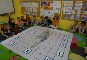 Grupa dzieci siedzi wokół rozłożonej na dywanie macie do kodowania. Na macie ułożony jest zegar z tabliczek. Dziecwyznka leży na macie ułożony w literkę I wskazując tym samym godzinę 12.